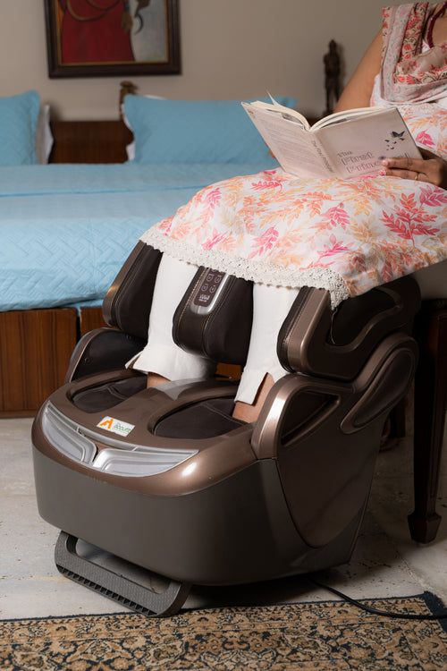 A brown and black foot massager placed on a bed in a bedroom, with a person sitting on the bed and reading a book while wearing white socks.
